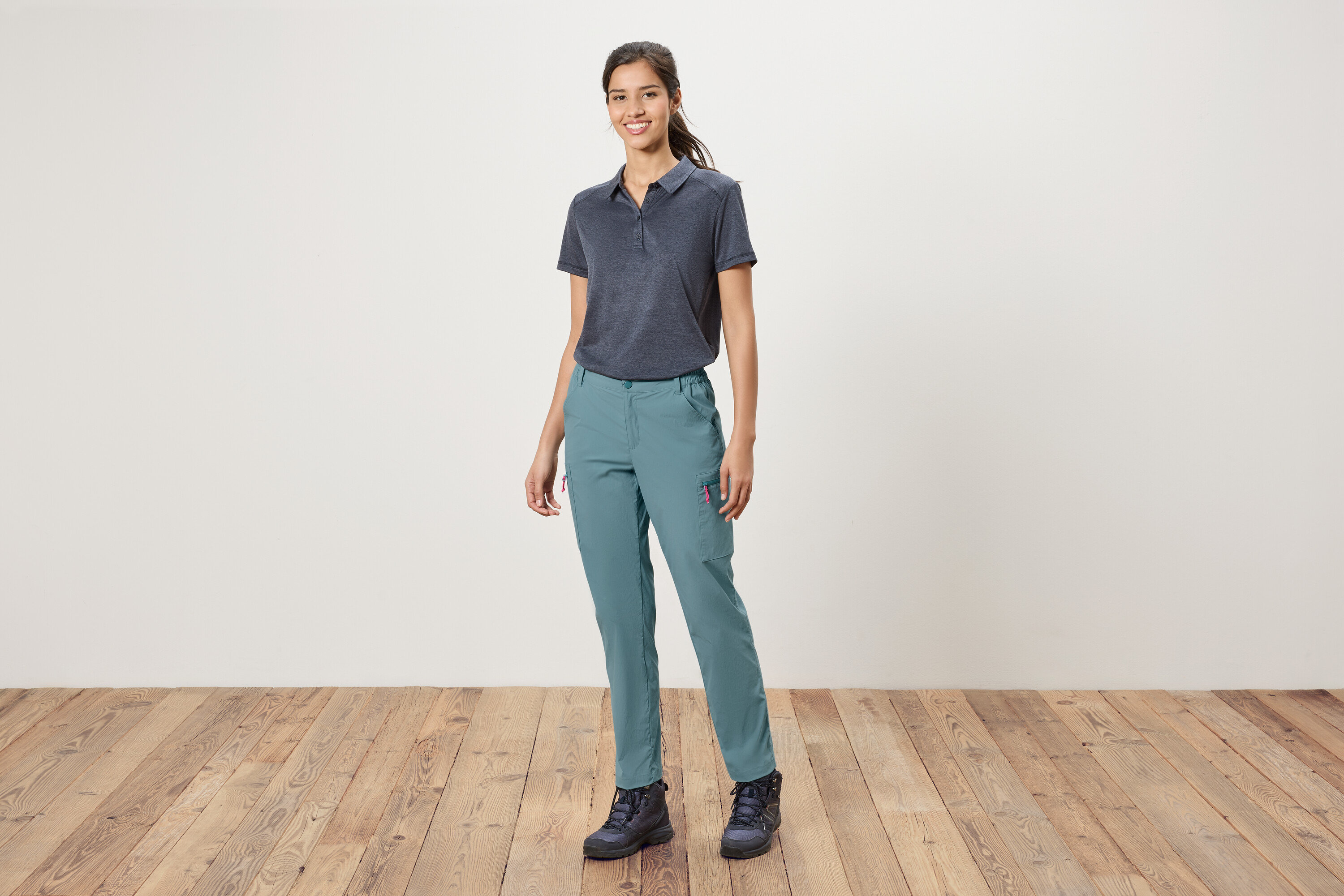 Žena stojí pred bielou stenou a má na sebe modré polotričko, modré nohavice a turistické topánky.