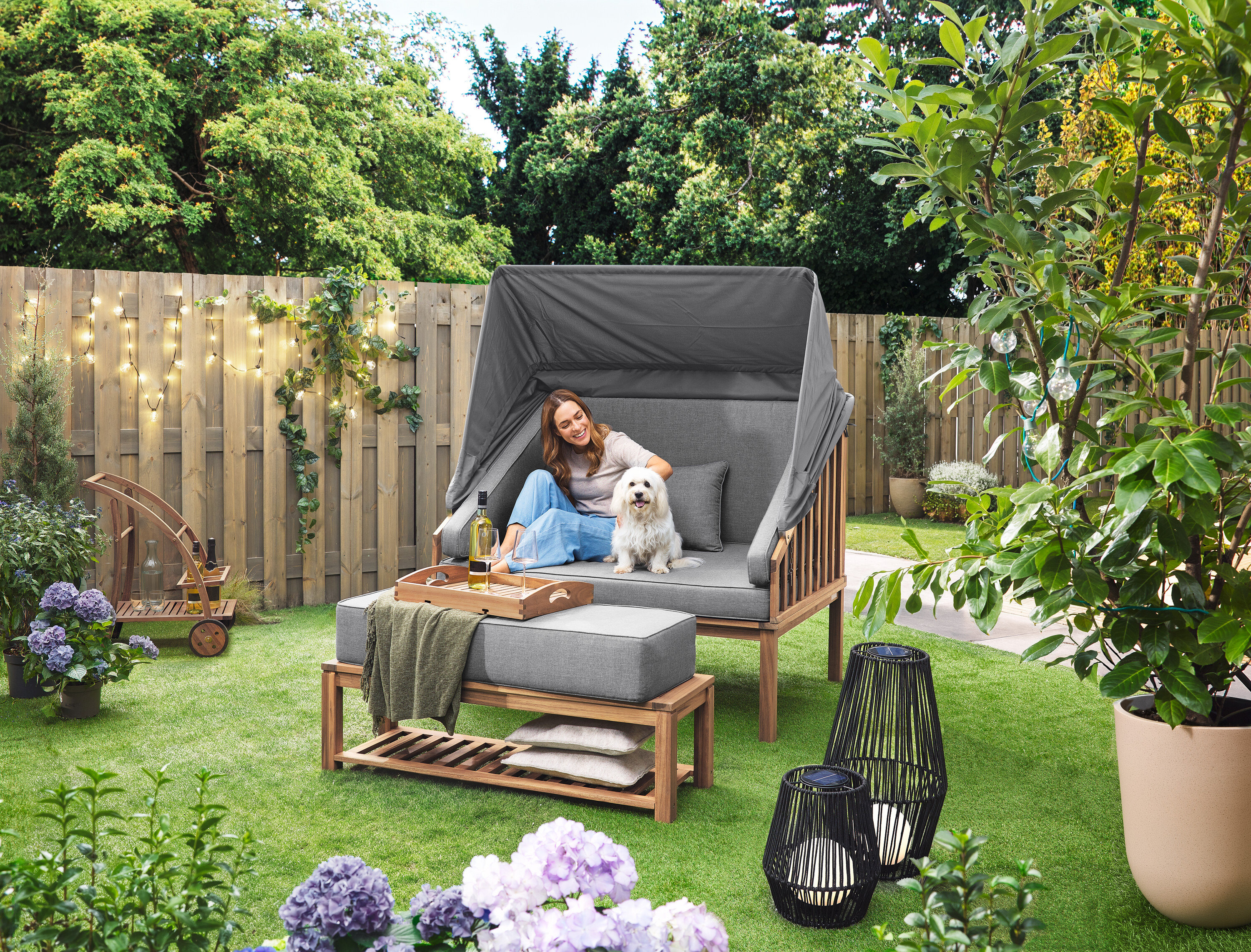 Žena sedí so psom v plážovom koši Premium Lounge-Strandkorb mit Sonnenschutz-Dach. Na trávniku stojí solárny lampáš s textilnou šnúrkou, veľký a solárny lampáš s textilnou šnúrkou, malý. V pozadí je barový vozík s odnímateľnou táckou. Na plote visí LED-Outdoor-Lichterkette mit Netzadapter.