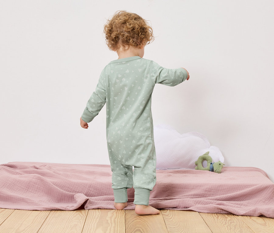 Malé dieťa stojí chrbtom k fotoaparátu na posteli s ružovou prikrývkou a zelenou hračkou.