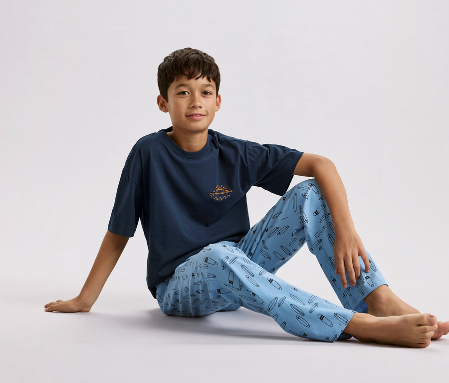 Chlapec sedí na zemi, má na sebe tmavomodré tričko s malým slnkom a vlnami a svetlomodré pyžamové nohavice so vzorom surfových dosiek.