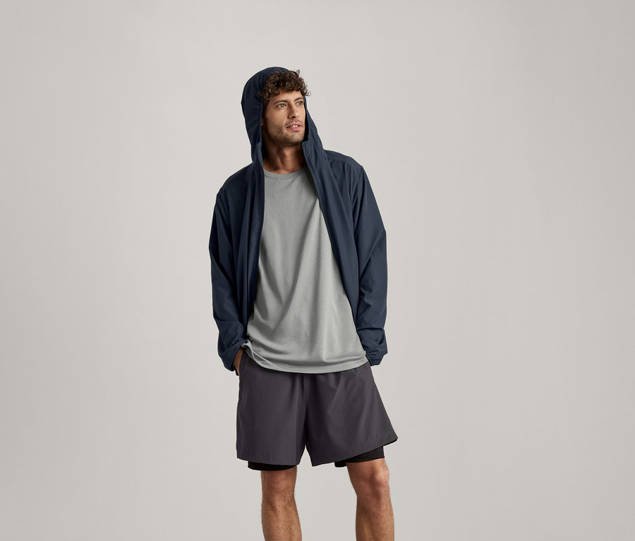 Muž má na sebe svetlosivé melírované športové tričko, tmavomodrú bundu s kapucňou a šortky.