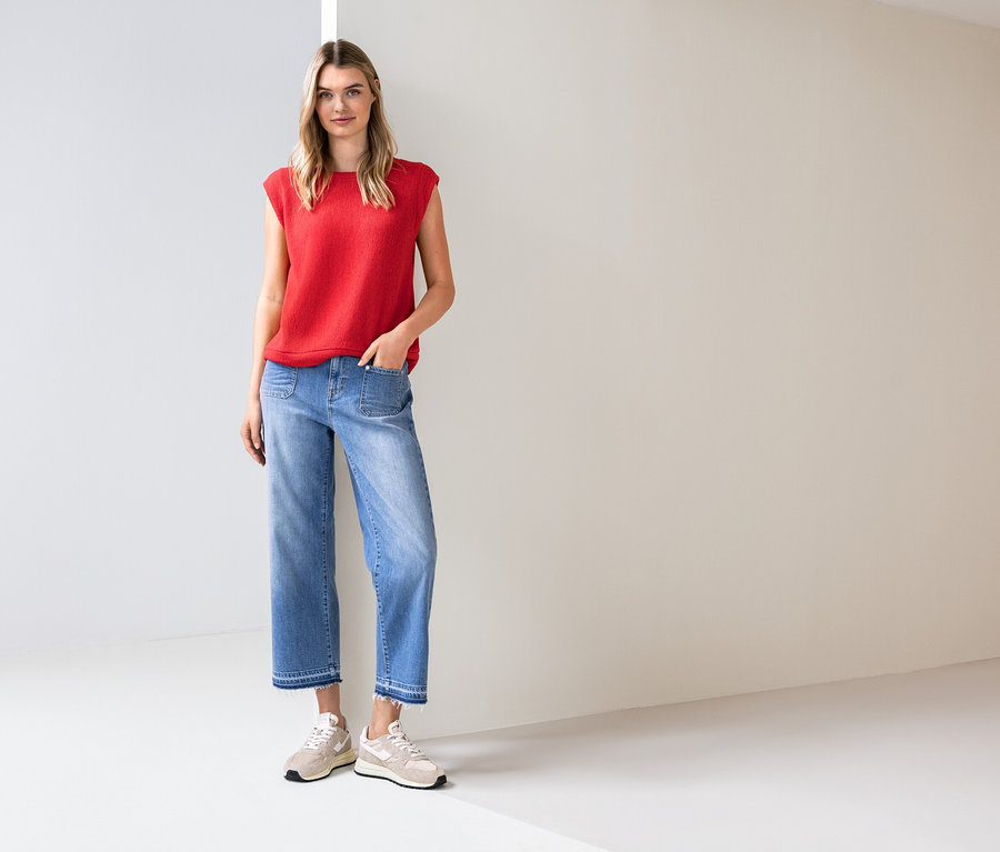 Žena stojí v svetlej miestnosti, má na sebe červený top, modré džínsy a tenisky.