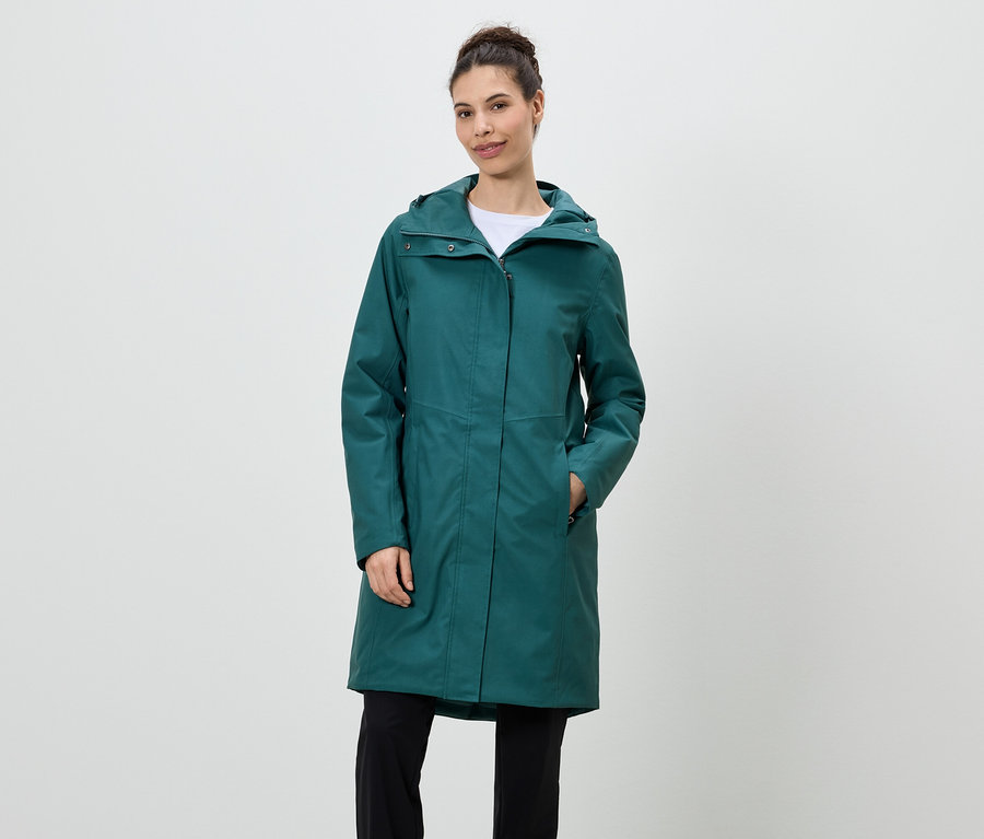 Žena stojí a má na sebe smaragdovozelený pršiplášť s kapucňou a čierne nohavice.