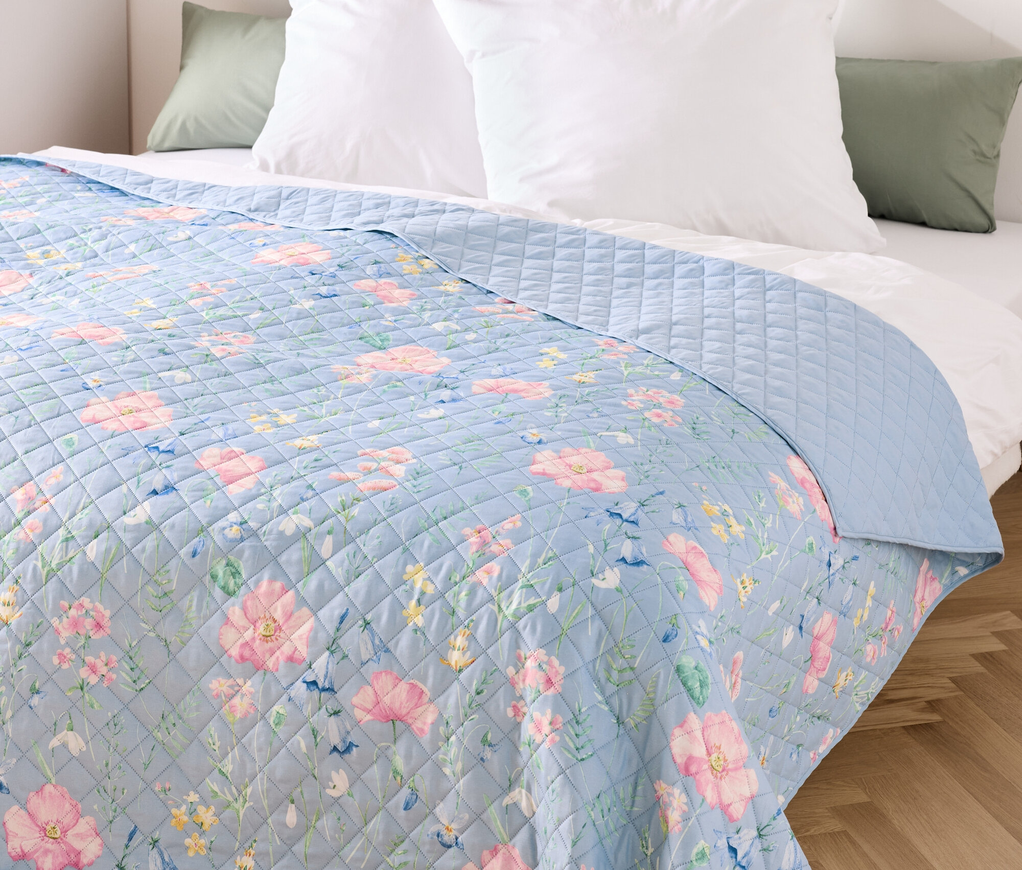 Modrý prehoz s kvetinovým vzorom na posteli s bielymi vankúšmi.