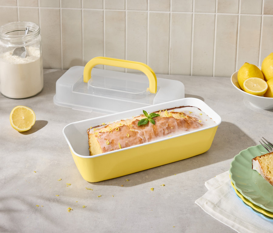 Citrónový koláč v žltej forme s citrónmi a polevou, nakrájaný na tanieri s vidličkou.