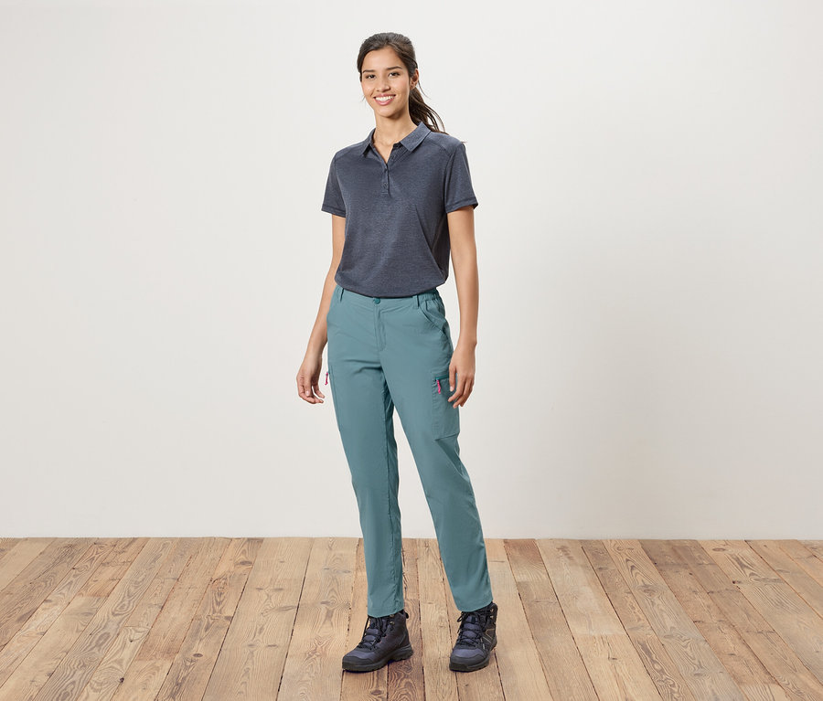 Žena stojí pred bielou stenou a má na sebe modré polotričko, modré nohavice a turistické topánky.