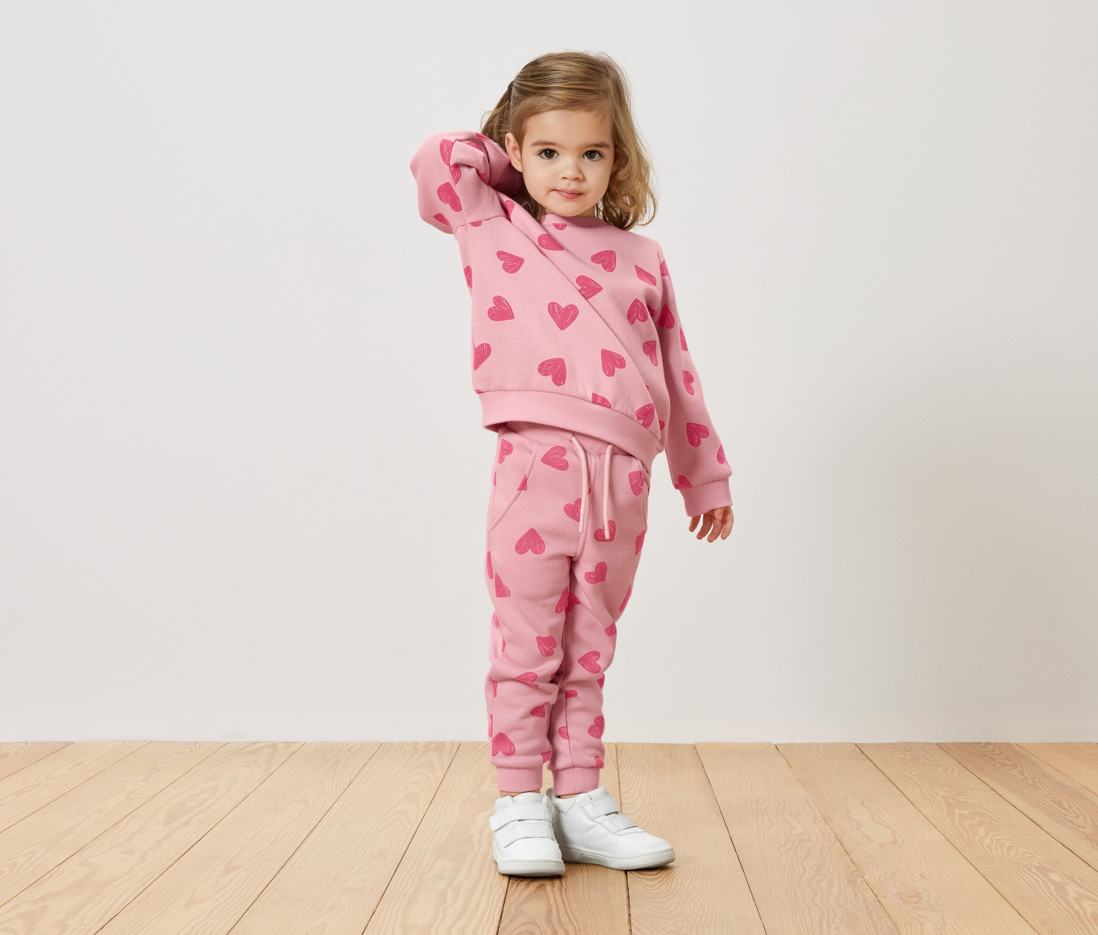 Dievča s blond vlasmi má na sebe ružový sveter a nohavice so vzorom srdca a biele tenisky.