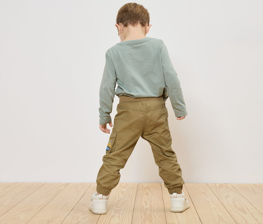 Dieťa stojí chrbtom k fotoaparátu, má na sebe zelené tričko s dlhým rukávom, khaki nohavice a biele tenisky.