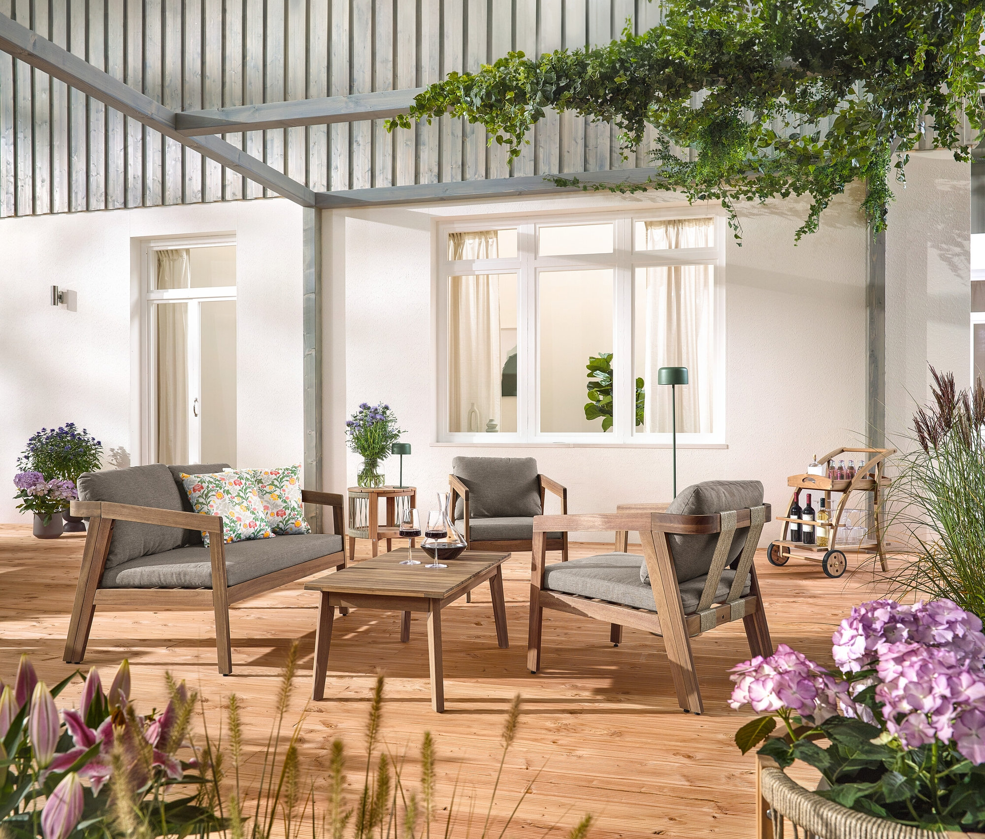 Na drevenej terase stojí 4-dielna záhradná sedacia súprava s textilným výpletom, s 2 vonkajšími vankúšmi s kvetinovým vzorom, barový vozík s odnímateľnou podnosom a záhradný príručný stolík s odnímateľnou doskou, Ø cca 50 cm. Vidieť aj solárnu nabíjaciu stolnú lampu, džungľovo zelenú a solárnu nabíjaciu stojacu lampu, džungľovo zelenú.