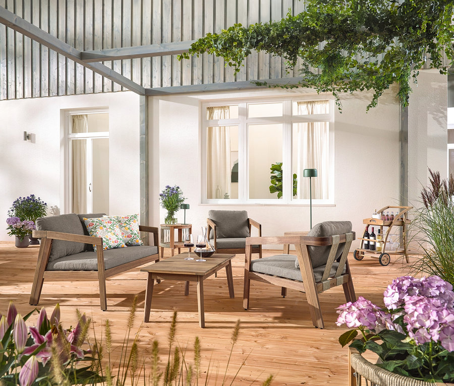 Na drevenej terase stojí 4-dielna záhradná sedacia súprava s textilným výpletom, s 2 vonkajšími vankúšmi s kvetinovým vzorom, barový vozík s odnímateľnou podnosom a záhradný príručný stolík s odnímateľnou doskou, Ø cca 50 cm. Vidieť aj solárnu nabíjaciu stolnú lampu, džungľovo zelenú a solárnu nabíjaciu stojacu lampu, džungľovo zelenú.