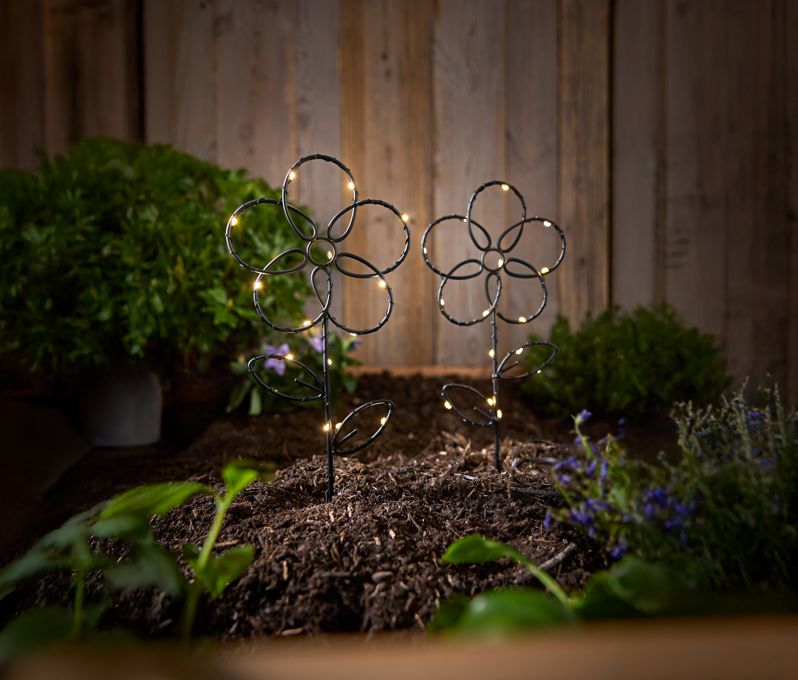 Dva čierne, kvetinové záhradné kolíky s LED svetlami stoja v zemi pred drevenou stenou.