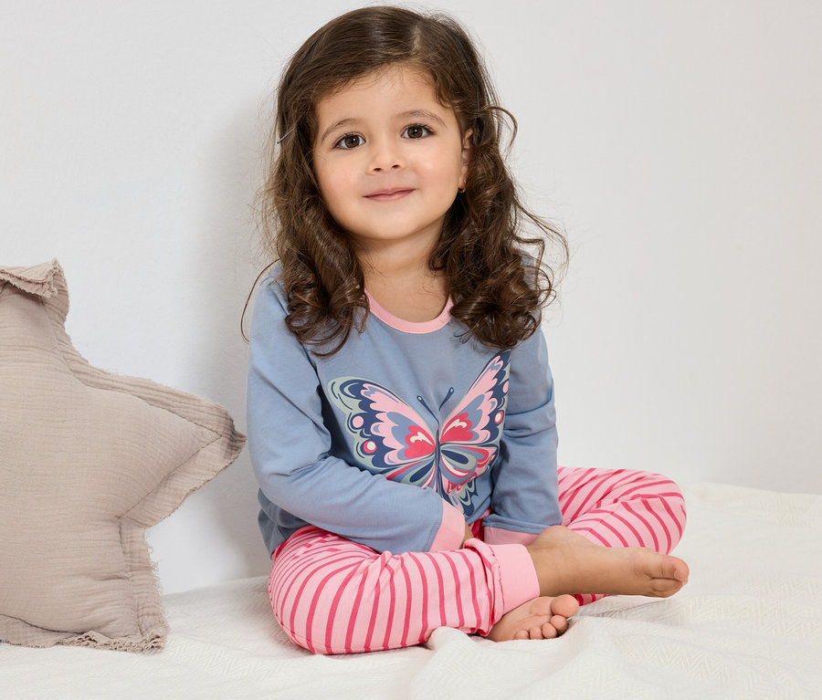 Dievča s kučeravými vlasmi sedí na posteli a má na sebe modré pyžamo s motýľom a ružové pruhované nohavice.