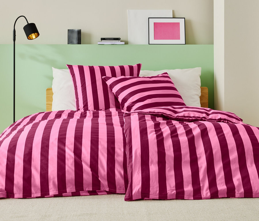 Posteľ s ružovo-vínovočervenými pruhovanými obliečkami, dvoma vankúšmi a stojacou lampou.