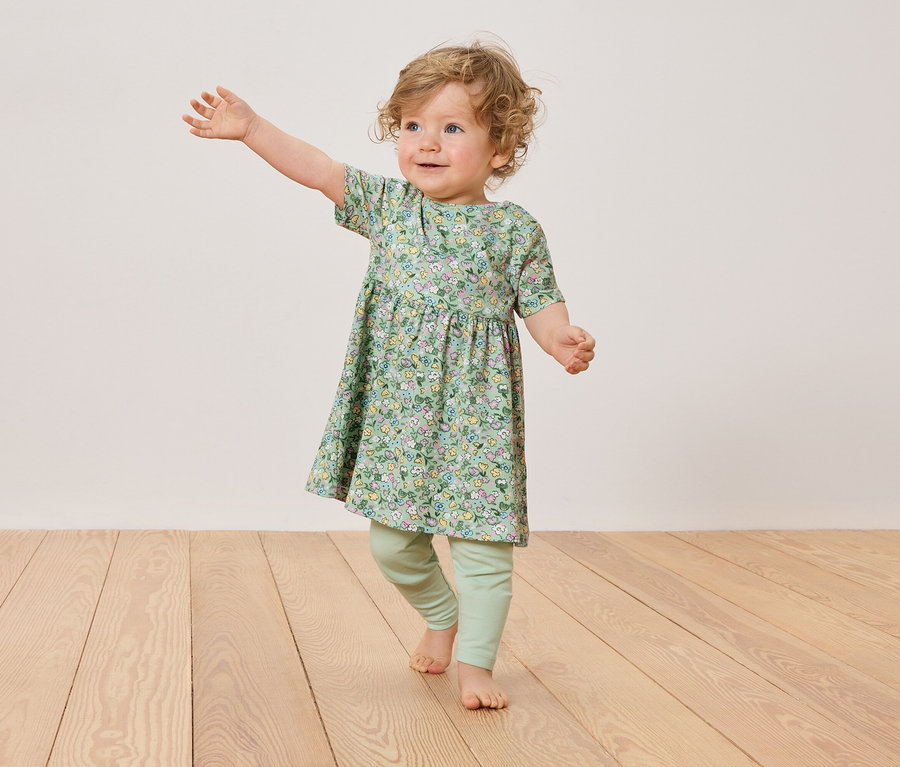 Malé dieťa s blond kučerami má na sebe kvetinové šaty a zelené legíny, stojí na drevenej podlahe.