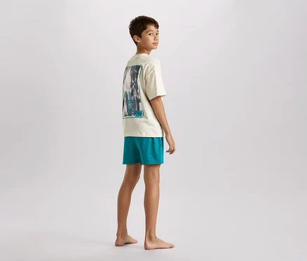 Mladý teenager stojí naboso a pozerá cez plece. Má na sebe krémové tričko s potlačou a tyrkysové šortky.