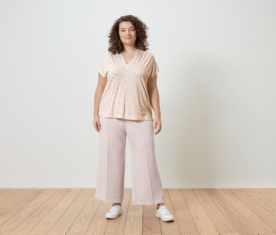 Žena s kučeravými vlasmi má na sebe bledoružovú vzorovanú blúzku, ružové culottes a biele tenisky pred bielou stenou.