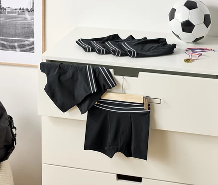 Na komode ležia čierne boxerky s bielymi pruhmi, futbalová lopta a medaila. Na vešiaku visia športové šortky.
