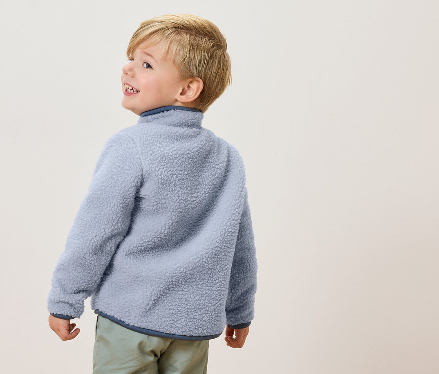 Malý chlapec s blond vlasmi má na sebe svetlomodrú fleecovú bundu a svetlozelené nohavice a pozerá sa nabok.