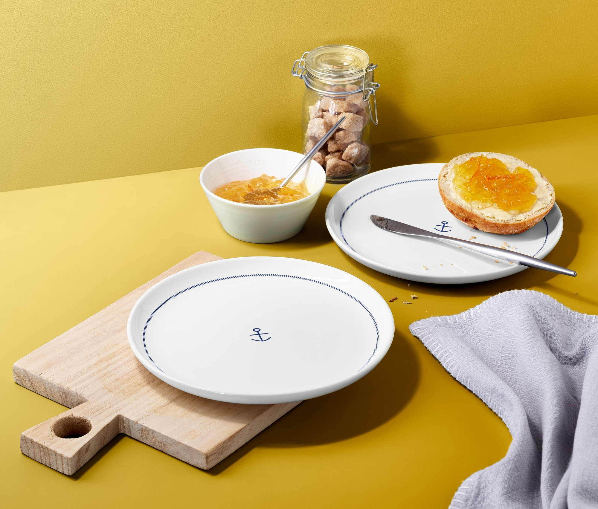 Dva taniere s motívom kotvy na drevenej doske a s chlebom s marmeládou na žltom pozadí.