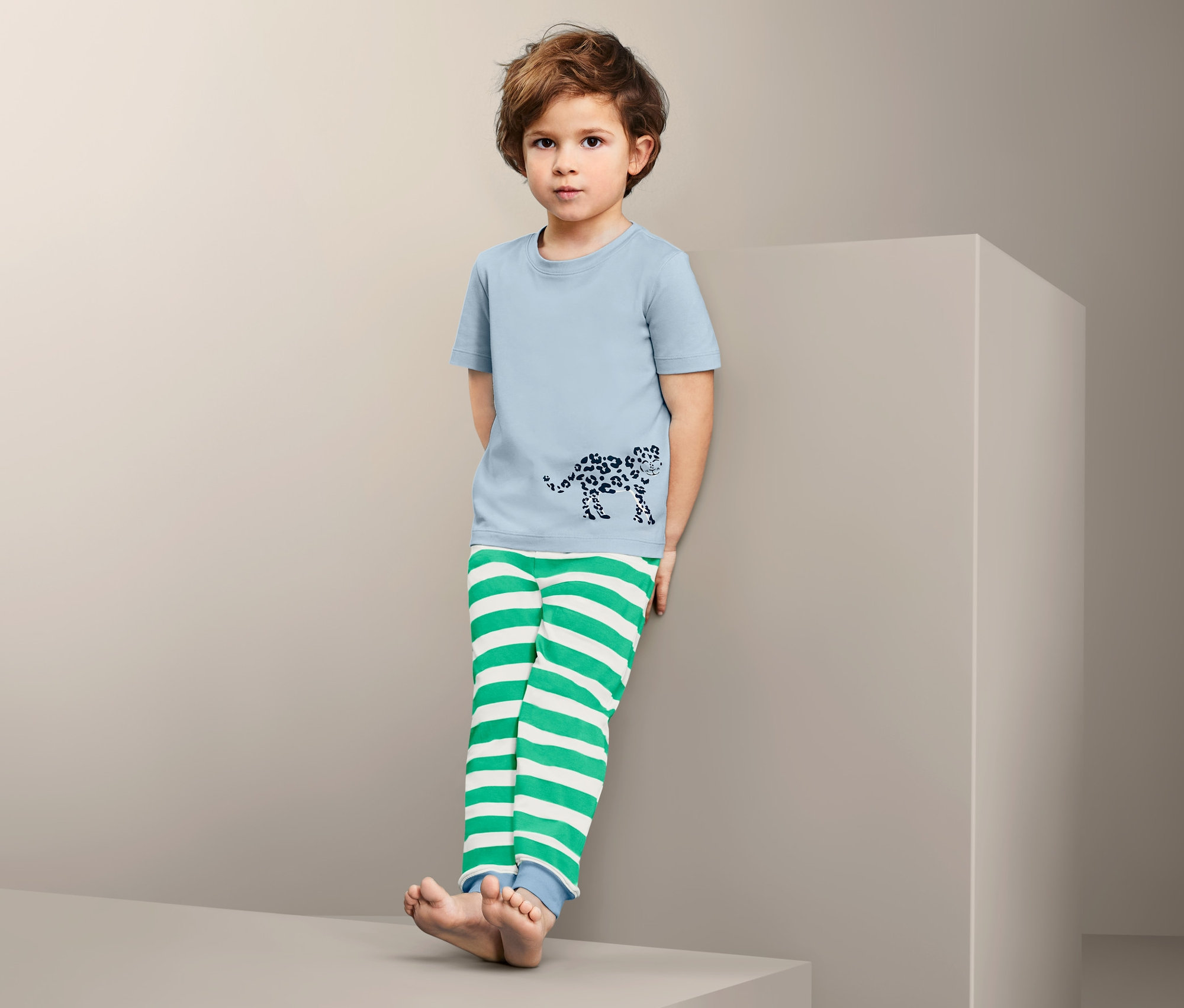 Chlapec v bavlnenom pyžame s modrým tričkom s potlačou leoparda a zeleno-bielymi pruhovanými nohavicami stojí opretý o stenu.