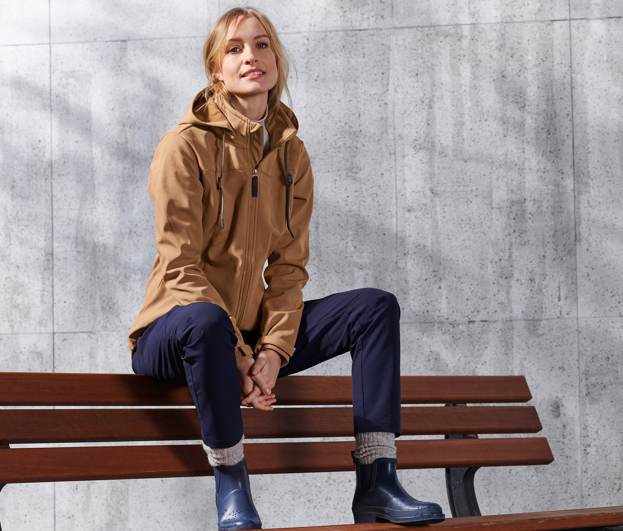 Blondínka sedí na lavičke a má na sebe koňakovú softshellovú bundu.