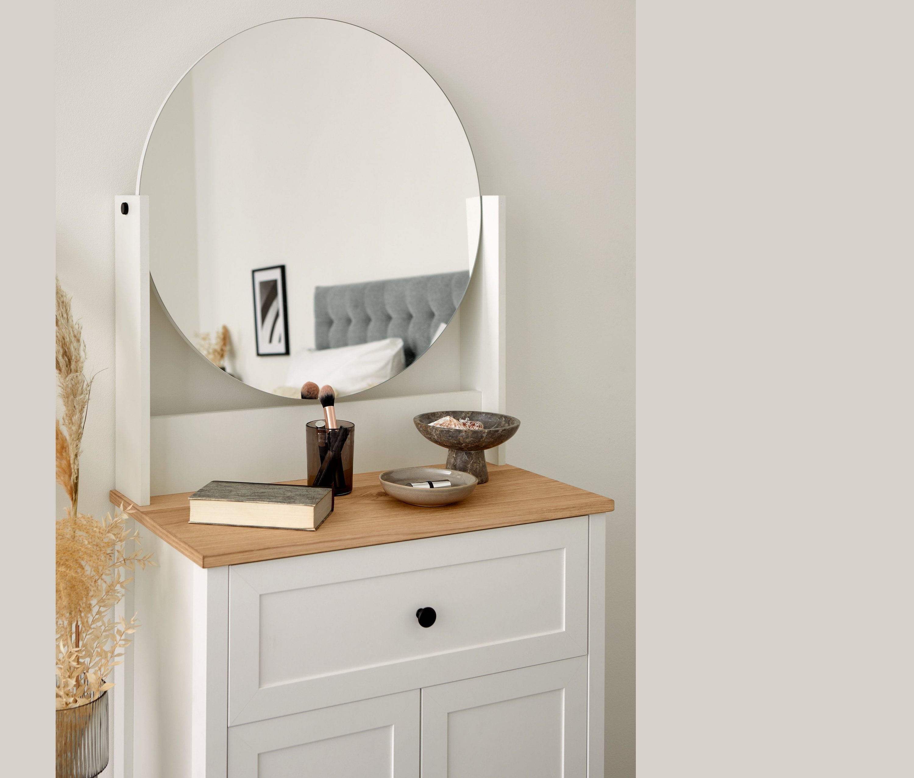 Toaletný stolík s okrúhlym zrkadlom, knihou, štetcami a ozdobnými miskami. V zrkadle sa odráža posteľ.