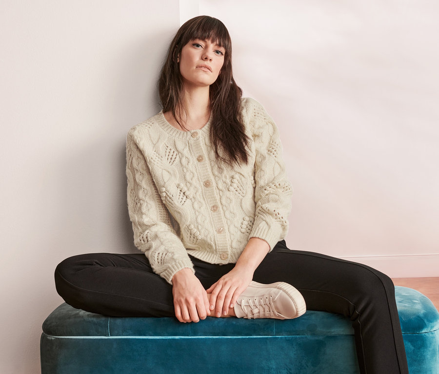 Žena sedí s prekríženými nohami na modrom zamatovom otomane a má na sebe kardigán z hrubej pleteniny.