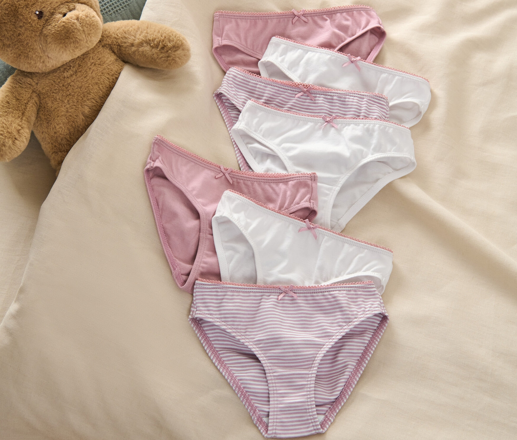 Sedem dievčenských nohavičiek leží na posteli vedľa plyšového medvedíka.