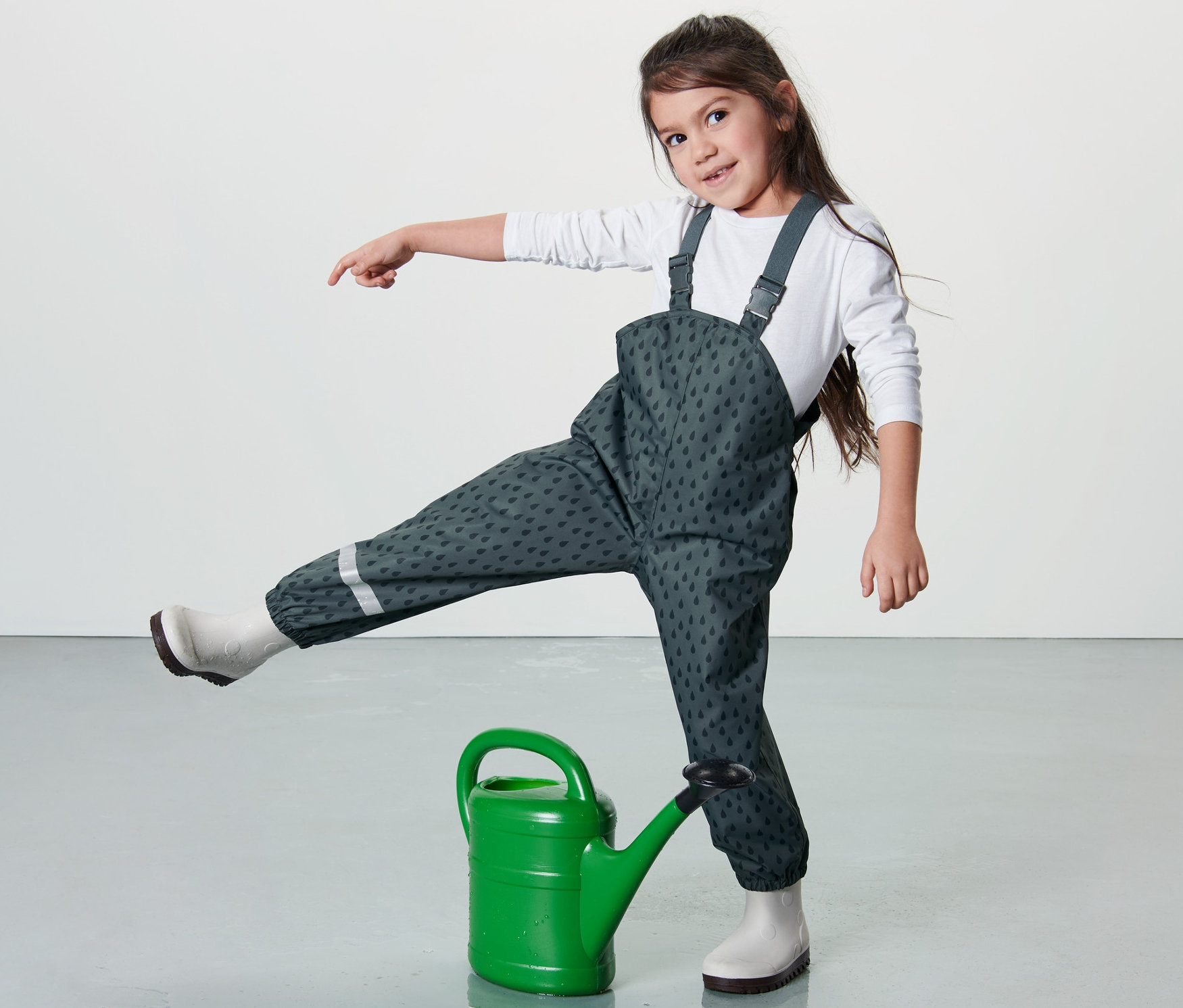 Dievča stojí jednou nohou na zelenej kanve a má na sebe detské nohavice do dažďa, bridlicovomodré.