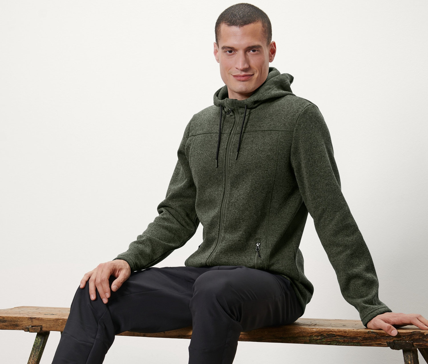 Muž sedí na lavičke a má na sebe bundu z pleteného flaušu.