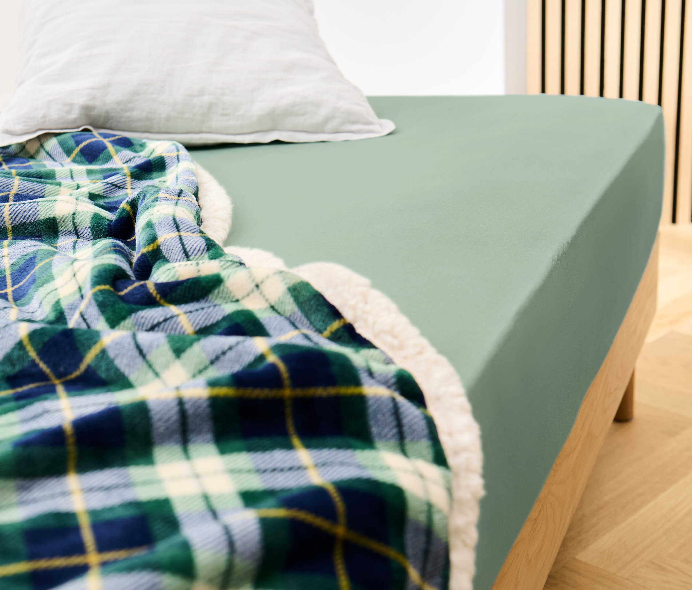 Posteľ s džersejovou plachtou šalviovej farby a kockovanou dekou.
