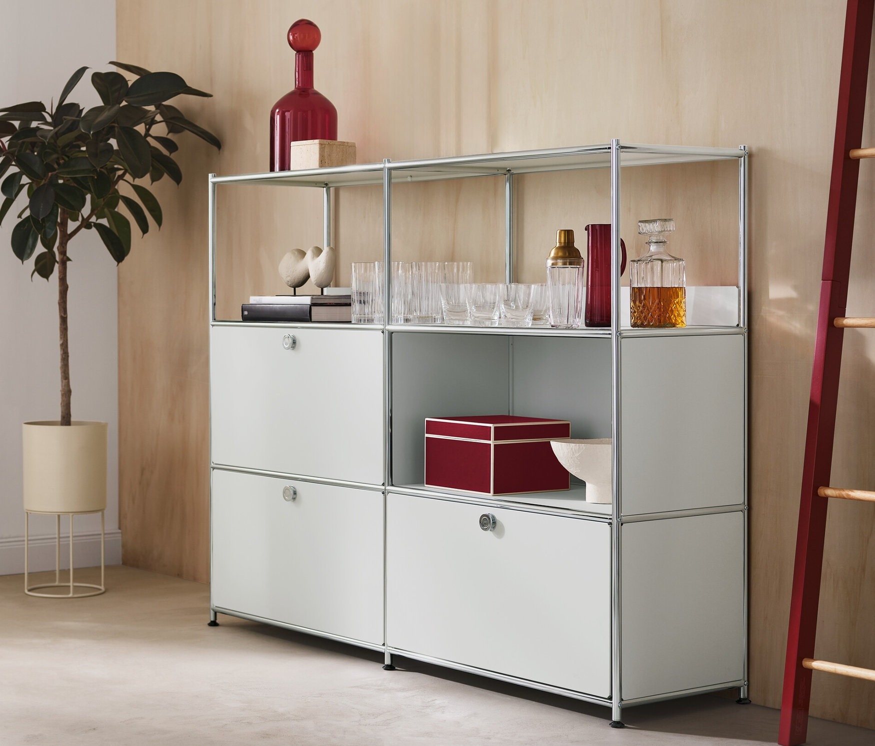 V miestnosti je okrúhly kvetináč s rastlinou, kovová komoda »CN3« s 2 výklopnými dvierkami a 1 zásuvkou a dekoračný rebrík s červenými bočnými prvkami.