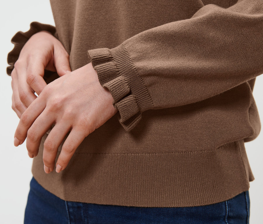 Osoba má na sebe hnedý pulóver z jemného úpletu s detailom v podobe volánikov a modré džínsy.