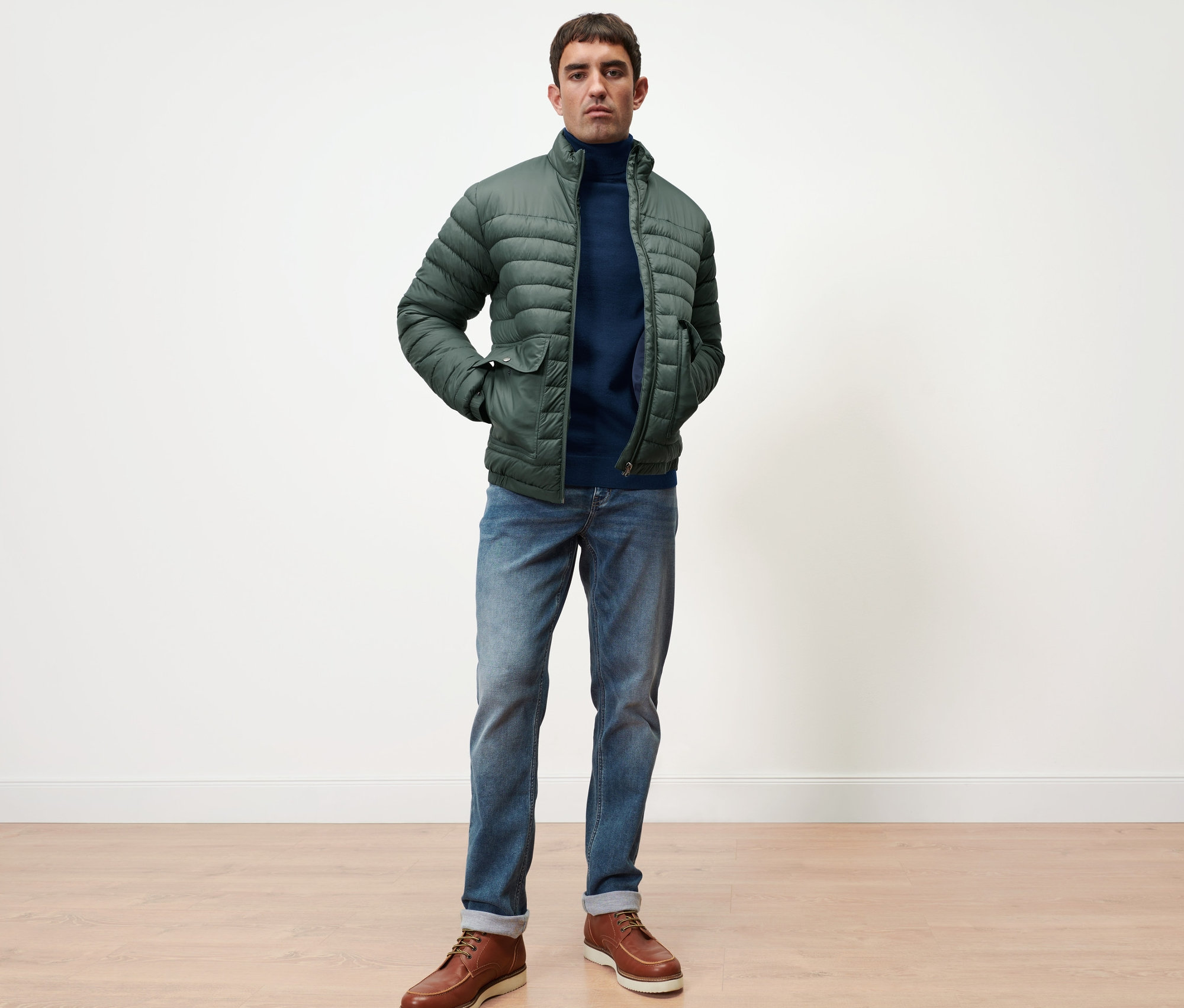 Muž stojí a má na sebe námornícky modrý pulóver z merino vlny a zelenú prešívanú bundu.
