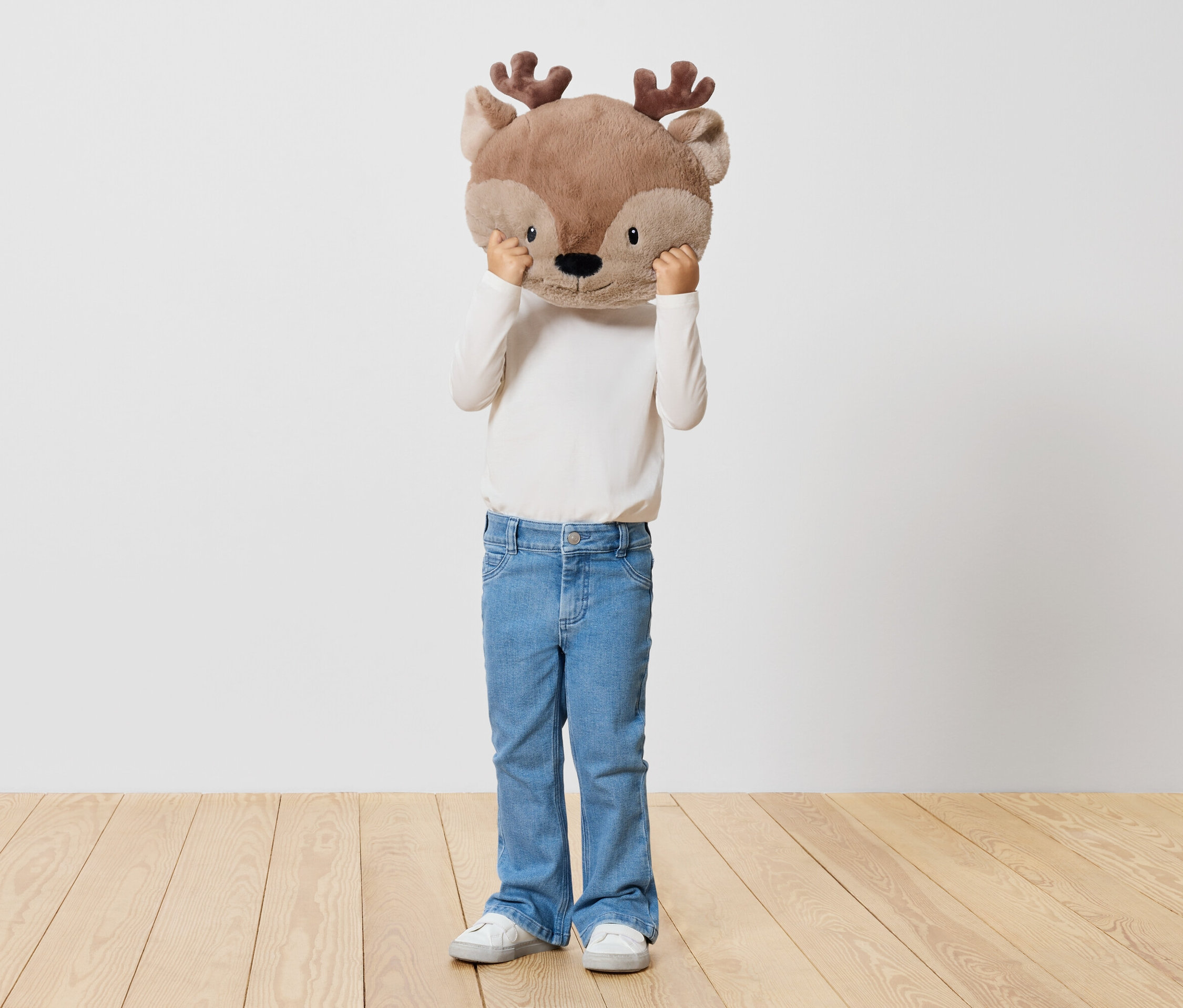 Dieťa drží plyšovú hračku jeleňa pred tvárou, stojí na drevenej podlahe pred bielou stenou.