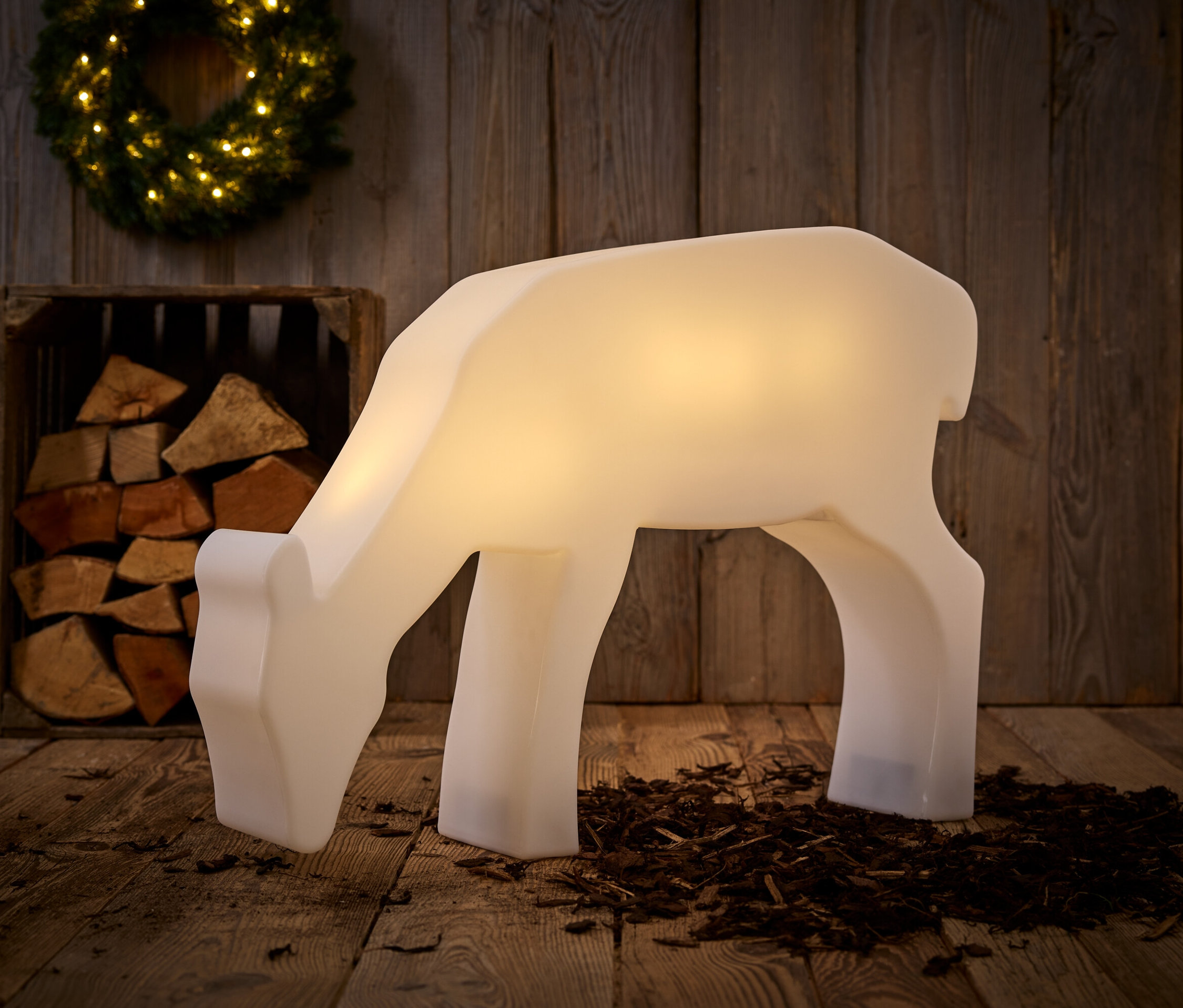 Svietiaca biela dekoratívna socha jeleňa stojí na drevenej podlahe, v pozadí je drevená stena, drevená debna s polenami a veniec.