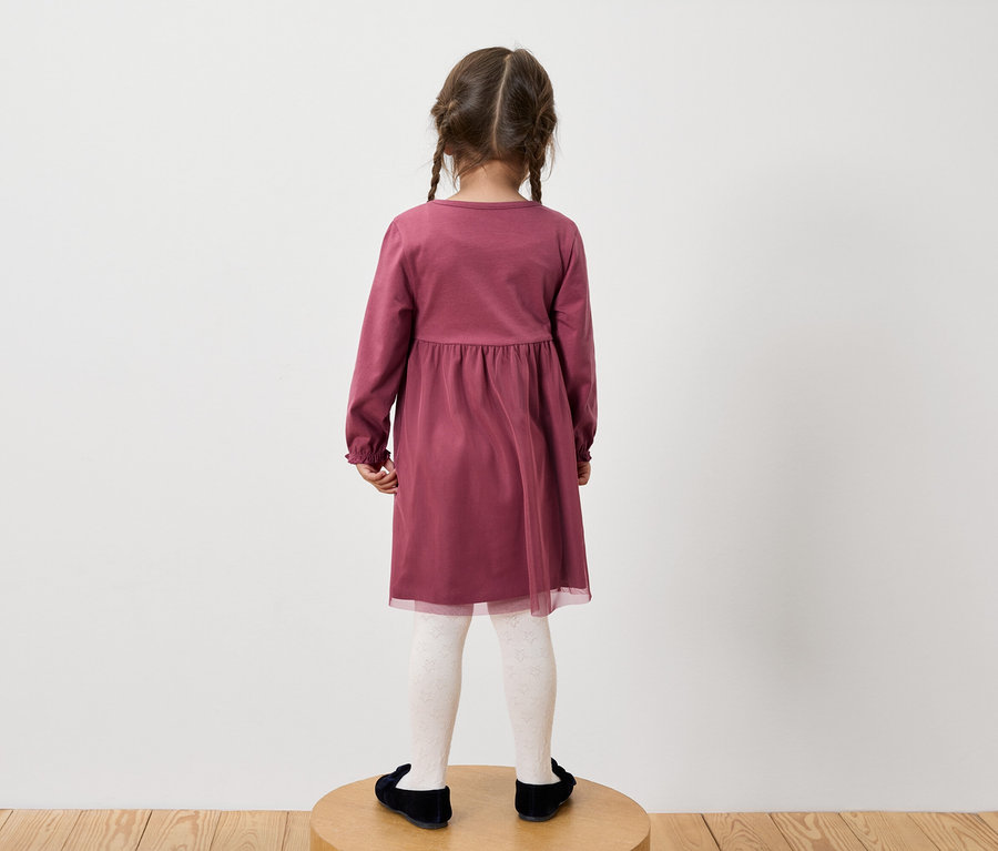 Dievča v bordových šatách s dlhým rukávom, bielych vzorovaných pančuchách a čiernych topánkach stojí chrbtom k fotoaparátu.