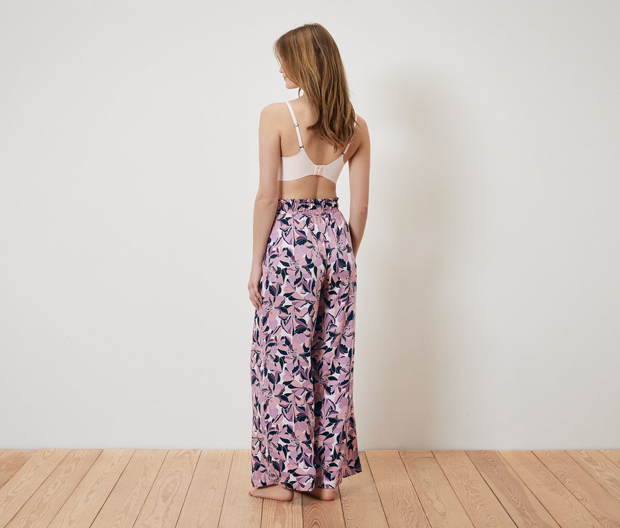 Žena chrbtom k fotoaparátu má na sebe ružovú podprsenku a široké nohavice s kvetinovým vzorom.