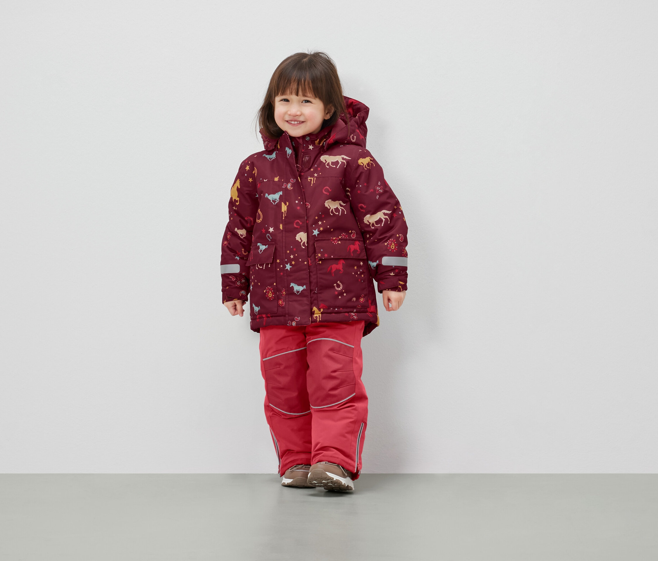 Dievča stojí a má na sebe zimnú bundu s kapucňou s potlačou koní a červené otepľovačky.