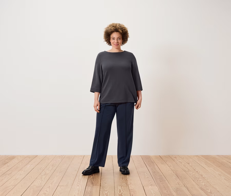 Žena s afro účesom stojí na drevenej podlahe a má na sebe sivú blúzku a tmavomodré nohavice.