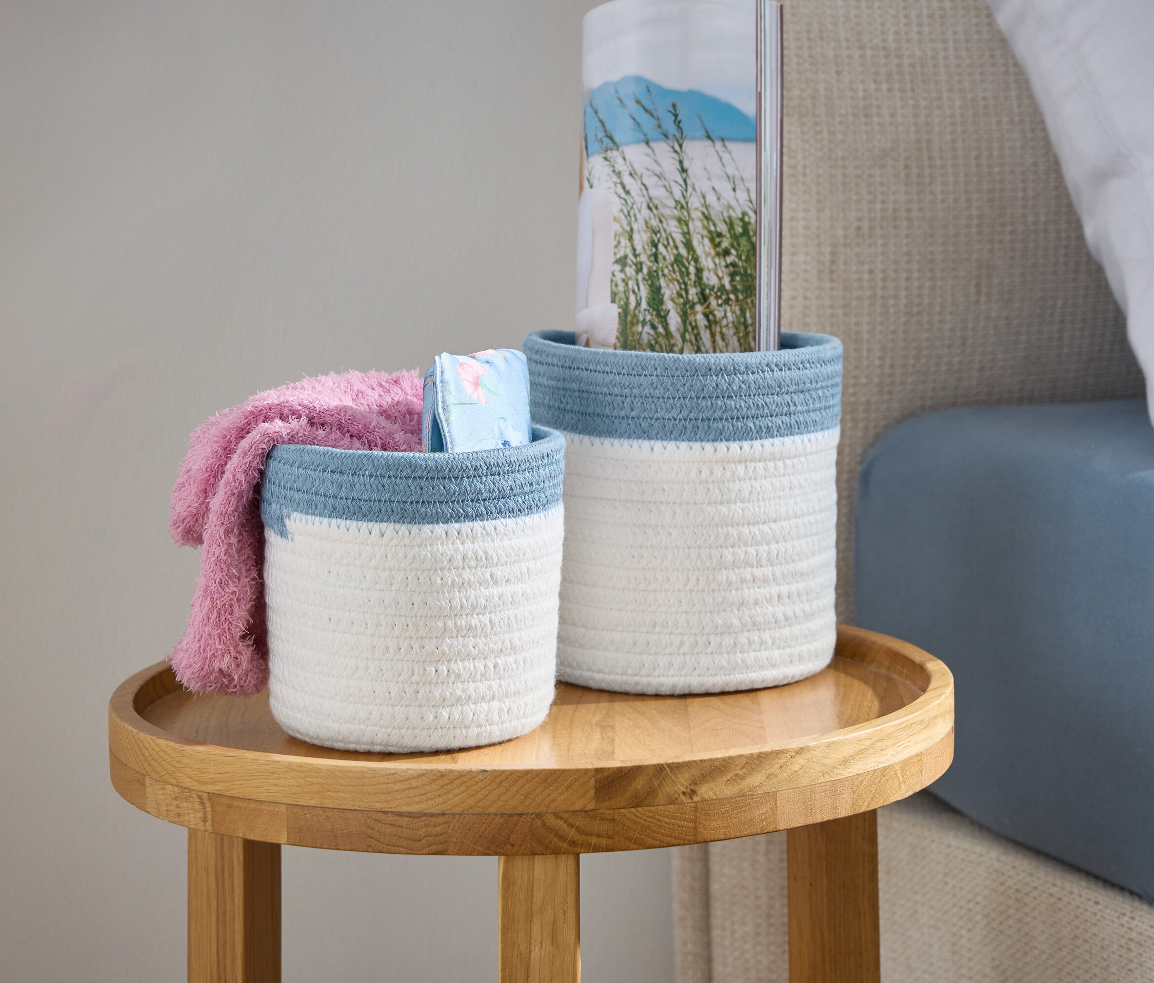 Dva biele pletené košíky s modrým lemom stoja na drevenom stolíku vedľa postele.