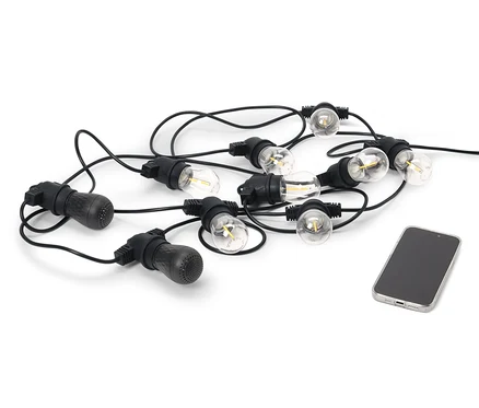 Čierna outdoorová LED svetelná reťaz s 2 Bluetooth® reproduktormi leží vedľa mobilného telefónu na bielom pozadí.