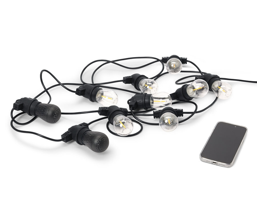 Čierna outdoorová LED svetelná reťaz s 2 Bluetooth® reproduktormi leží vedľa mobilného telefónu na bielom pozadí.