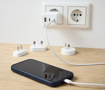 Biely USB cestovný adaptér zapojený do zásuvky nabíja mobilný telefón. Na stole sú ďalšie adaptéry.