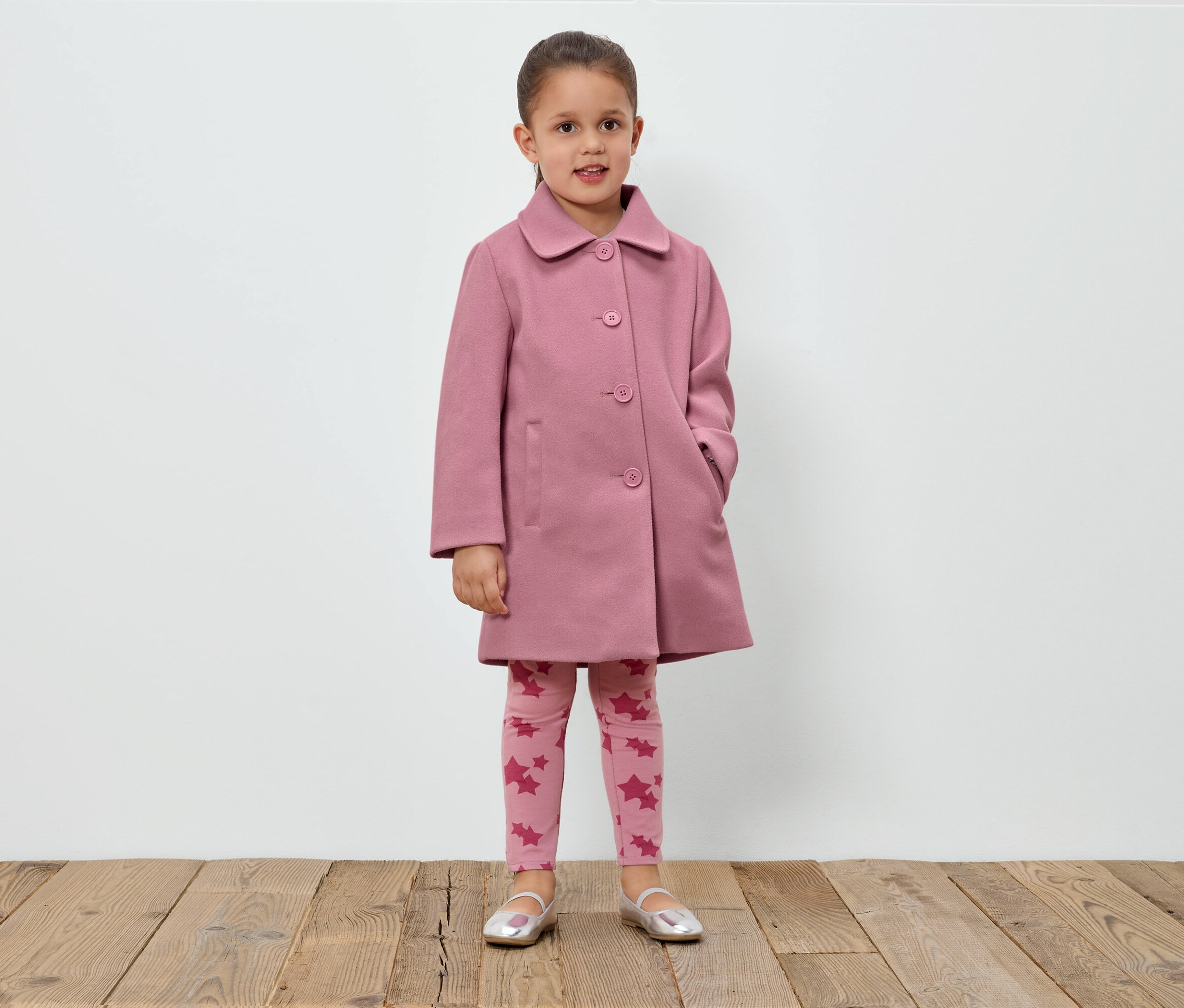 Dievča stojí pred bielou stenou a má na sebe ružový kabát, legíny s hviezdami a strieborné topánky.