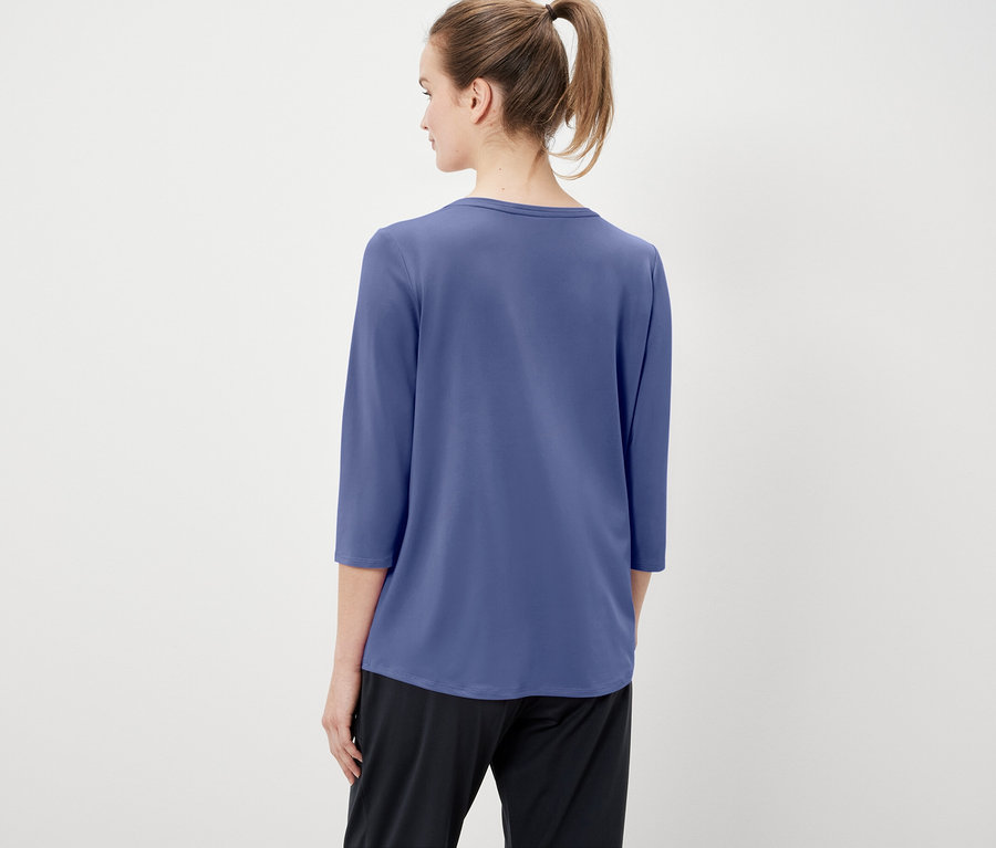 Pohľad zozadu na ženu s vlasmi stiahnutými do copu, oblečenú v modrom tričku s trojštvrťovými rukávmi a čiernych nohaviciach.