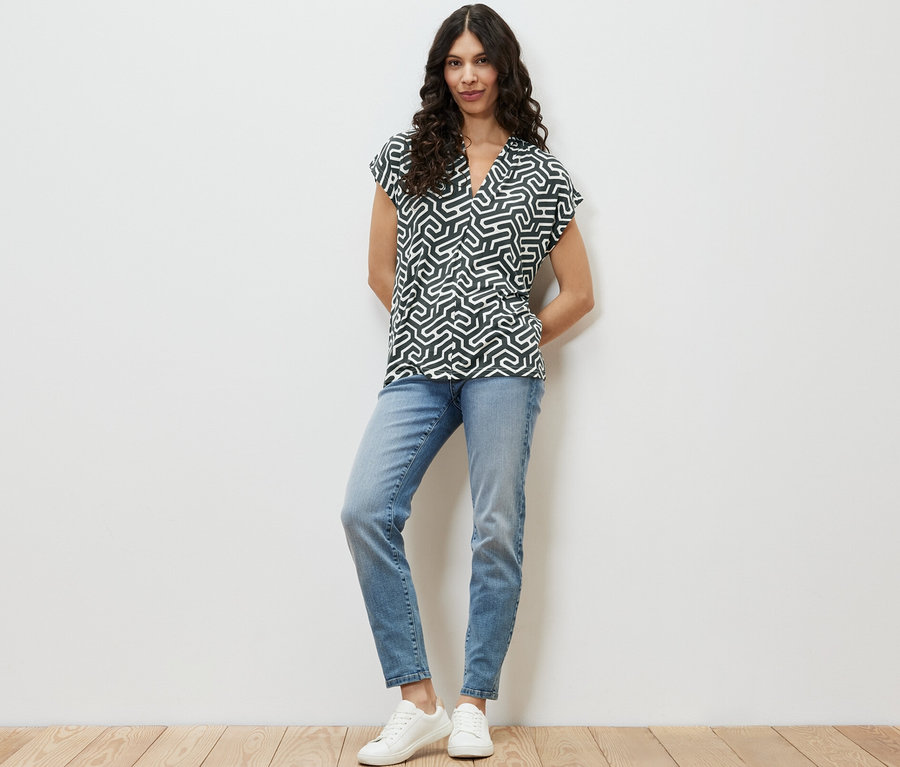 Žena stojí a má na sebe zelené blúzkové tričko s výstrihom do V, modré džínsy a biele tenisky.