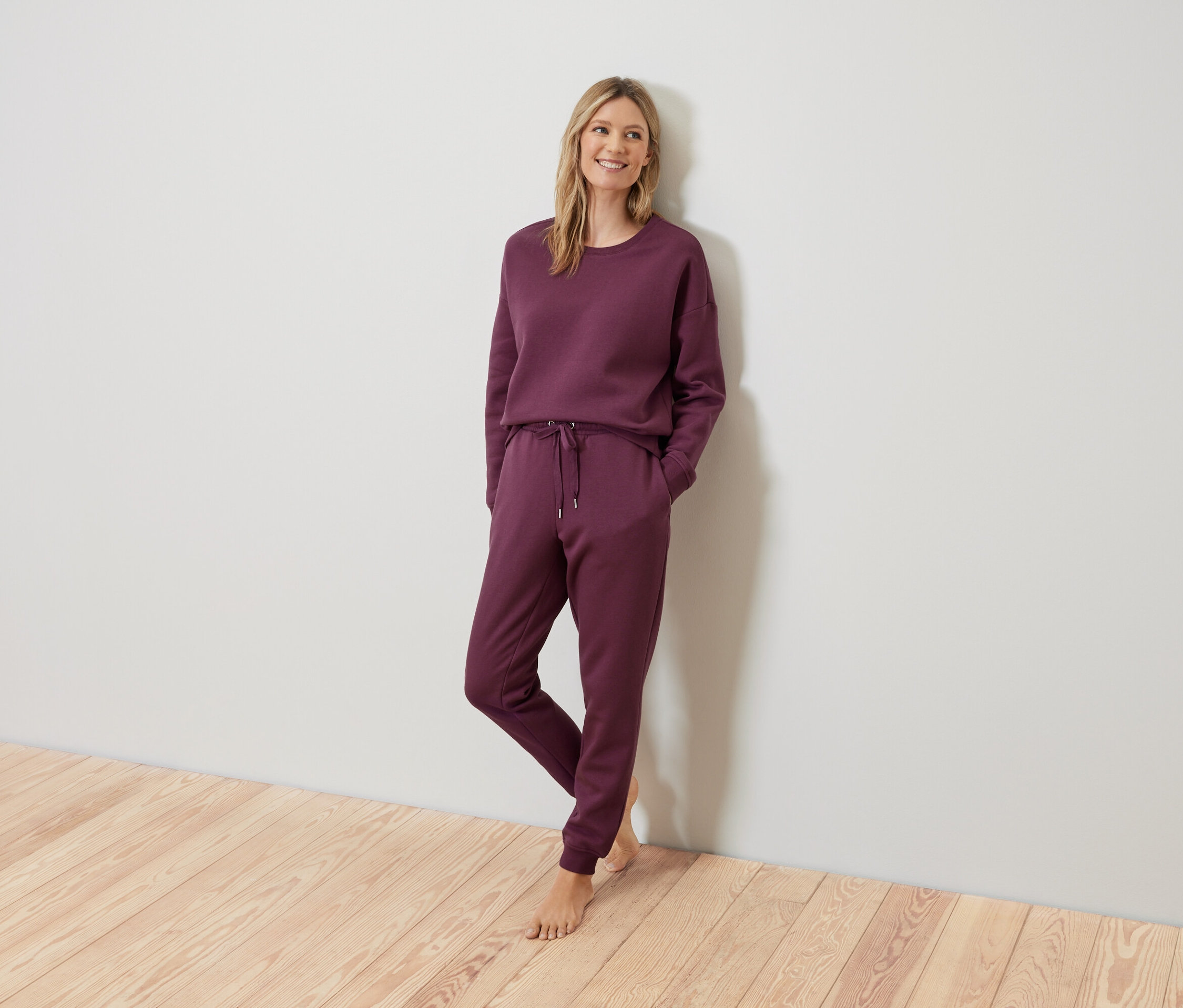 Blondínka pózuje v bordovej teplákovej súprave pri stene.