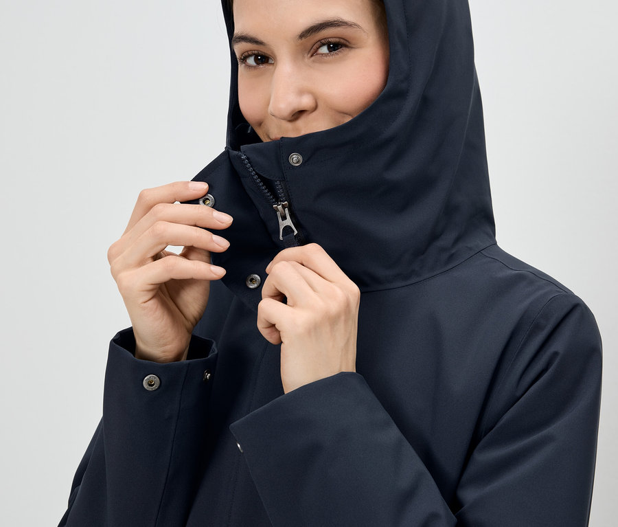 Žena si zapína zips na tmavomodrej bunde s kapucňou.