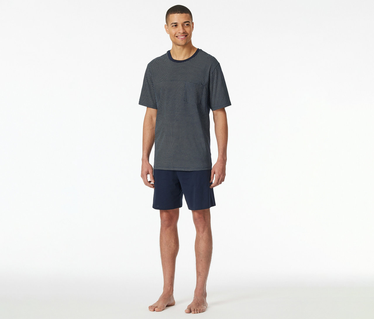 Muž stojí naboso na bielom pozadí v krátkom pyžamovom sete SCHIESSER pozostávajúcom z tmavomodrého trička s krátkym rukávom a bodkami a šortiek.